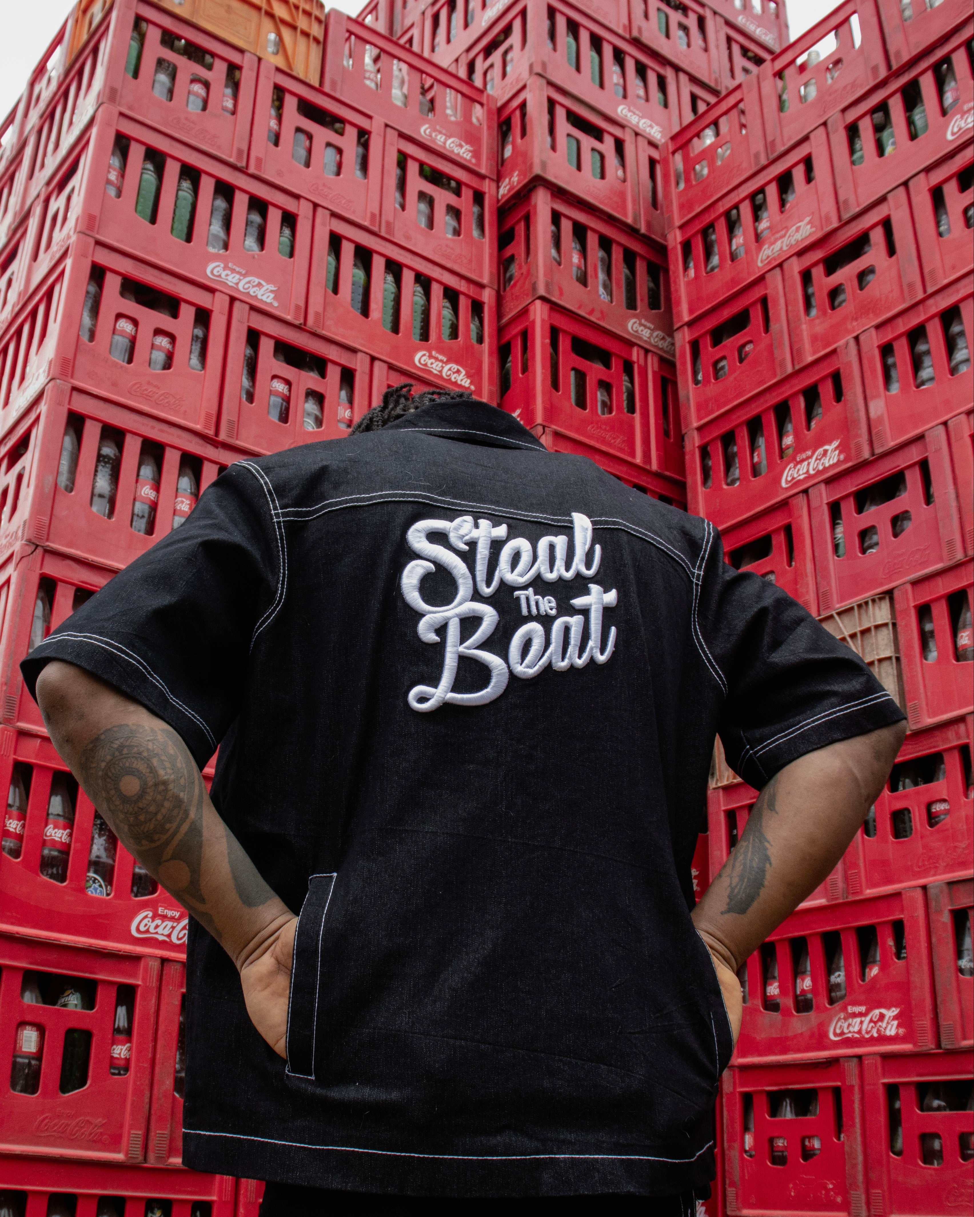 Person wearing a black jacket with 'Steal The Beat' on the back, standing in front of stacked red crates.