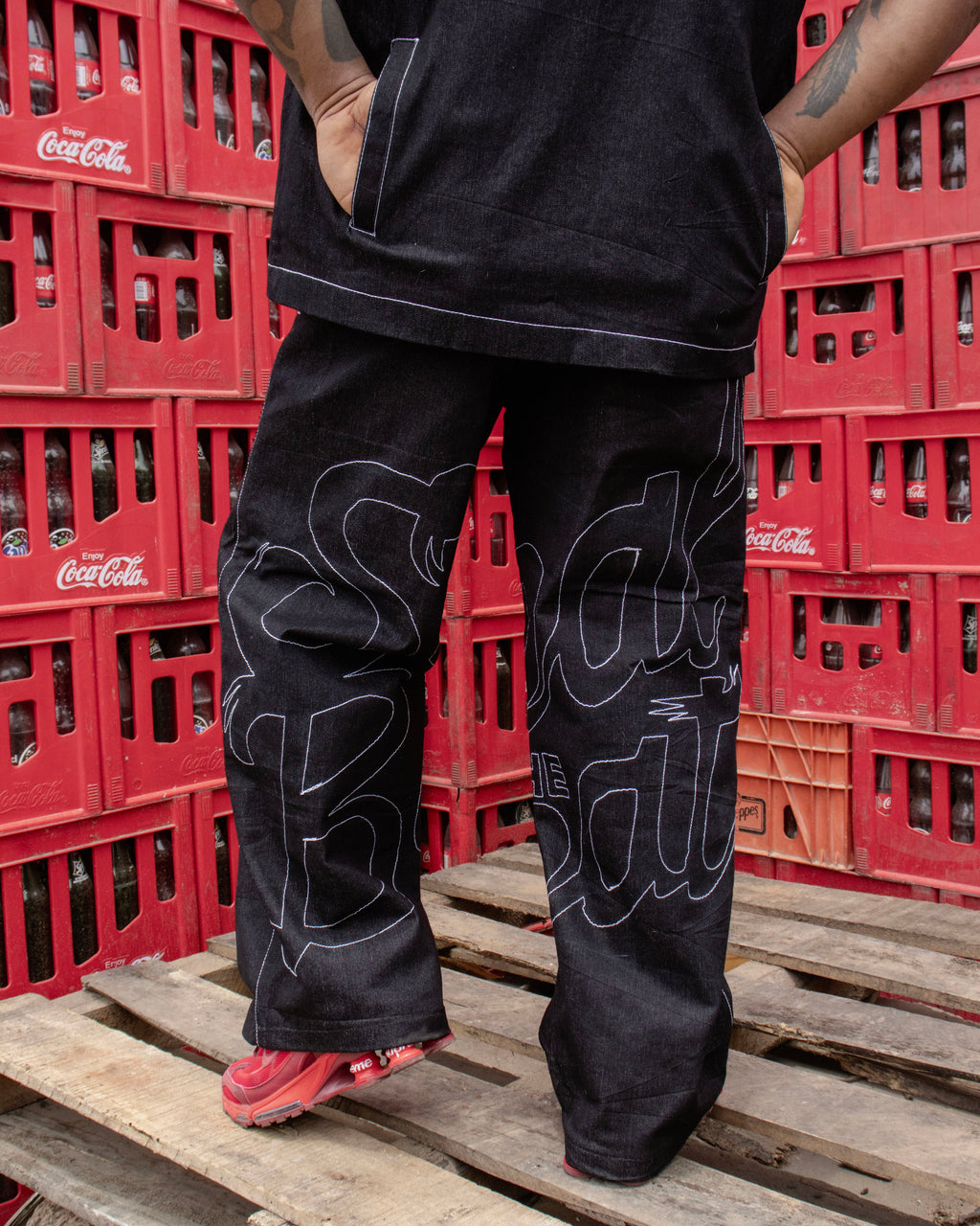 Person wearing black pants with white patterns standing on wooden pallets with red crates in the background.